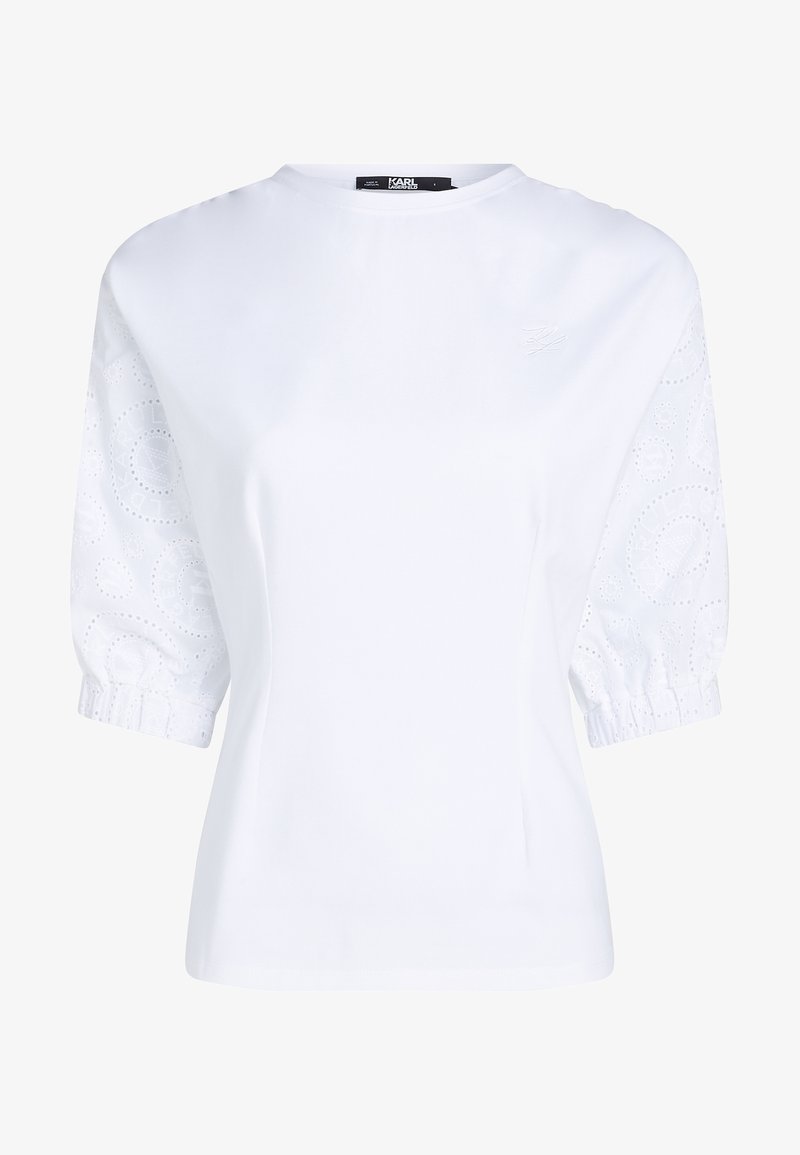 Blouse blanche à manches courtes avec manches bouffantes en dentelle à jours et col rond, avec une broderie discrète du logo sur la poitrine.