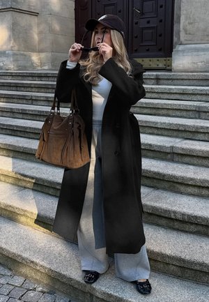Une femme portant une casquette noire et un long manteau tient des lunettes de soleil près de son visage, sur des marches en pierre, et porte un grand sac à main marron.