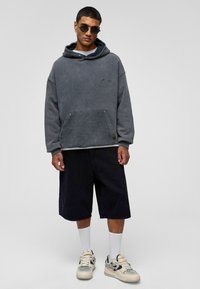 Grijze fleece hoodie met een buidelzak, gecombineerd met zwarte shorts. Het model draagt grijze sneakers en witte crew-sokken, met ronde zonnebrillen.