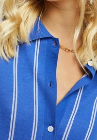 Camisa de punto azul con rayas blancas, cuello abotonado y detalles de botones visibles. Presenta un corte holgado y una textura suave.