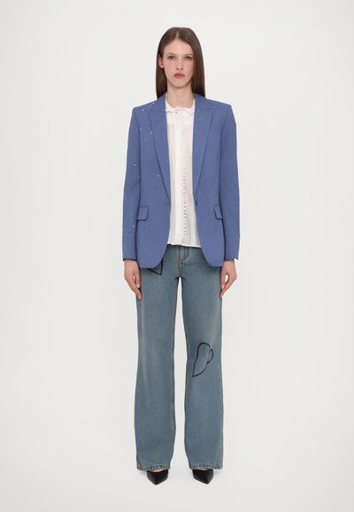 Blauwe blazer met versieringen, witte blouset met ruches en verontrustende lichtblauwe jeans met een hartvormige patch; zwarte puntige schoenen.