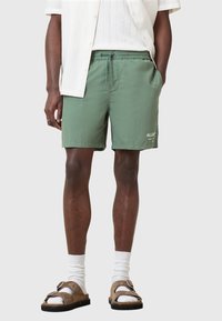 Shorts verdi con vita elastica e cordoncino, caratterizzati da un design semplice con un logo bianco. Il modello indossa sandali marroni e calze bianche.