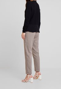 Schwarzes, strukturiertes Langarmoberteil, kombiniert mit hellbraunen und schwarzen karierten Hosen. Abgerundet wird das Outfit durch weiße, mit Riemchen versehene Sandaletten mit hohen Absätzen.