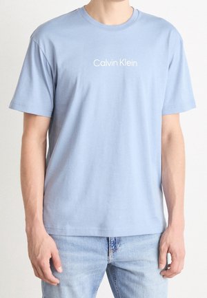 Muž ve světle modrém tričku Calvin Klein a světle modrých džínách stojí před jednobarevným světlým pozadím.