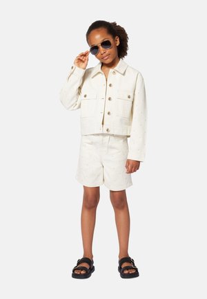 Veste en denim blanc raccourcie avec de grandes poches et des boutons dorés, associée à un short blanc assorti présentant des motifs de points dorés. Sandales noires et lunettes de soleil.
