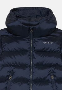 Marineblå pufferjakke med høy krage, frontglidelås og to glidelåslommer på siden. Stoffet har en glatt, blank tekstur. GANT-logo på brystet.