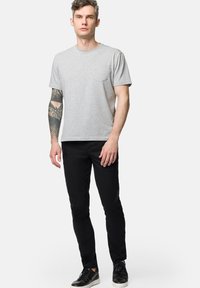 Szara bawełniana koszulka z kieszonką na piersi, zestawiona z czarnymi dopasowanymi spodniami i czarnymi sznurowanymi butami. Model ma tatuaż na lewym ramieniu.