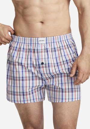 Man wearing white plaid boxer shorts with blue and red stripes, pulling waistband slightly outward showing elastic fit.