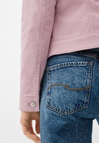 Chaqueta de cuero rosa claro con botones en los puños, combinada con jeans de mezclilla azul desgastados que presentan un destacado detalle en las costuras de los bolsillos.