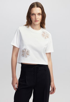 T-shirt bianca cropped con ricami floreali in beige e accenti neri davanti. Abbinato a pantaloni neri. Texture di cotone liscia.