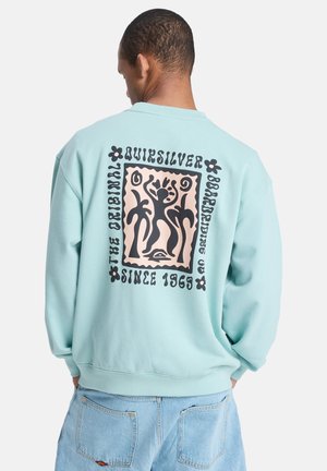 Lichtblauwe sweatshirt met zwart en beige grafisch ontwerp op de rug, met abstracte figuren, omringd door tekst en bloemaccenten.