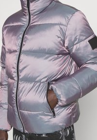 Nafukovací bunda v duhově světle fialové barvě, s vysokým límcem, zipovým zapínáním, bočními kapsami a logem na rukávu.
