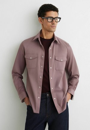 Muž s krátkymi kučeravými vlasmi a okuliarmi, oblečený v mauve košeli na gombíky cez tmavé tričko a tmavé džínsy, stojaci proti bielej stene s rukami v vreckách.