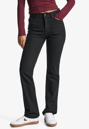 Person trägt schwarze Jeans mit geradem Bein, ein dunkelrotes Langarmshirt und weiße Sneaker mit braunen Sohlen und steht mit einer Hand in der Tasche.