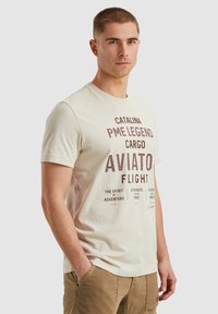 T-shirt en coton beige clair à manches courtes, avec des textes et des éléments graphiques maroon liés à l'aviation et à l'aventure.