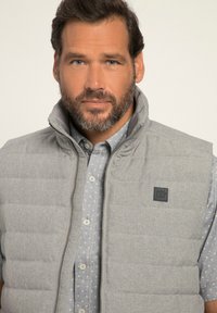 Gilet imbottito grigio con motivo a rombi, colletto alto, zip frontale e una piccola etichetta nera, indossato sopra una camicia blu con pois gialli.