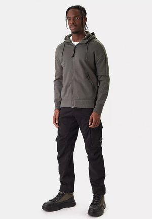 Uomo con capelli intrecciati che indossa una felpa grigia con cerniera, pantaloni cargo neri e sneakers nere alte, in piedi davanti a uno sfondo semplice.