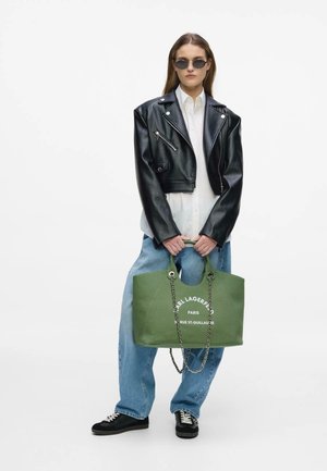Femeie purtând o jachetă de piele neagră, cămașă albă, blugi albaștri, ochelari de soare, ținând o geantă tote verde Karl Lagerfeld cu mânere din lanț.