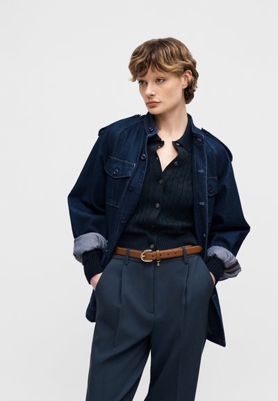 Person med kort, bølget hår iført mørk denimjakke over sort kabelstrikket sweater, stoppet ned i navybukser med brunt bælte, hænderne i lommerne.