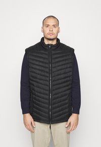 Gilet imbottito nero con colletto alto, caratterizzato da motivi a righe orizzontali, una zip frontale e tasche laterali. Indossato sopra una camicia blu navy.