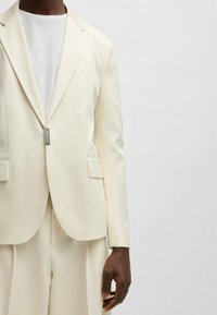 Cream blazer with sharp lapels, single button closure, two front pockets, and smooth texture. Worn over a white shirt.