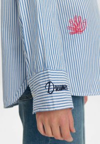 Gestreiftes blaues und weißes Hemd aus Baumwollstoff, mit einem roten bestickten Design und "Dreamer" in Marineblau am Bund.