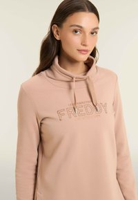 Haut de hoodie rose clair avec un col roulé haut, arborant un logo imprimé "FREDDY". Texture douce et design minimal, avec des cordons de serrage pour un ajustement.