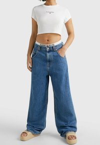 Krojované bílé tričko s potištěným logem, spárované s volnými modrými džíny a světle béžovými platformovými sandály. Džíny mají označený pás.