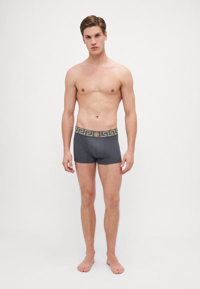 Boxer in cotone grigio con una cintura decorativa nera e oro che presenta un motivo geometrico. Il modello è in piedi a piedi nudi su uno sfondo bianco.