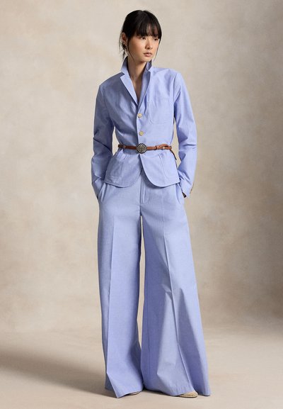 Γυναίκα που φοράει ανοιχτό μπλε, ραμμένο κοστούμι με φαρδιά παντελόνια και καφέ ζώνη, στέκεται με τα χέρια στις τσέπες μπροστά σε ουδέτερο φόντο.