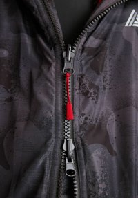 Jachetă neagră cu fermoar, cu un model de camuflaj, având un dispozitiv de deschidere roșu și accesorii metalice. Textură moale și material ușor.