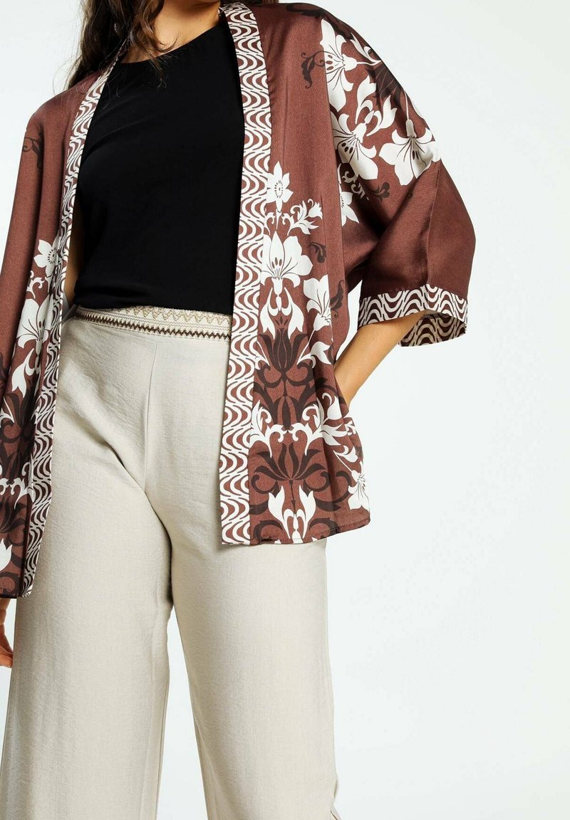 Femme portant un pantalon beige à jambes larges avec une ceinture brodée, un haut noir, et une veste kimono marron à motifs floraux avec des motifs blancs.