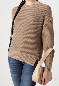 Personne portant un pull beige en maille épaisse et un jean bleu, tenant un petit sac à main beige avec une large bandoulière dans une main.