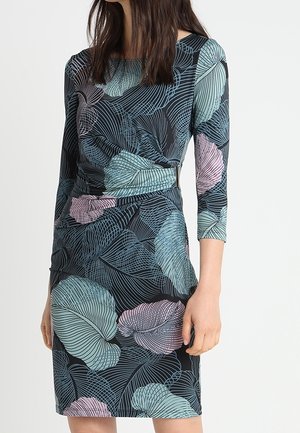 Frau trägt ein figurbetontes, knielanges Kleid mit dreiviertellangen Ärmeln und einem großen pastellfarbenen Blattmuster in Blau, Rosa und Schwarz.