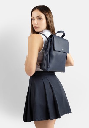 Giovane donna con top bianco senza maniche e gonna plissettata blu navy che porta uno zaino in pelle blu navy sulla schiena, guardando oltre la spalla.