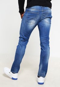 Pantalones vaqueros de mezclilla azul con un corte ajustado, lavado desgastado, cinco bolsillos y costuras en contraste, combinados con zapatillas blancas.