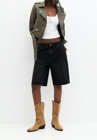 Shorts di denim neri con orlo sfrangiato, abbinati a una maglietta bianca attillata e un giubbotto di pelle verde oliva. Indossati con stivali da cowboy in suede color nocciola.