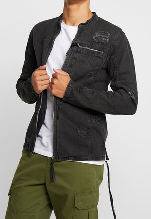 Personne portant une veste noire zippée et usée par-dessus une chemise blanche et un pantalon cargo vert olive sur un fond uni.