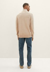 Suéter beige claro con cuello alto, puños y dobladillo acanalados; combinado con jeans azules y zapatos beige, visto desde atrás sobre un fondo neutral.