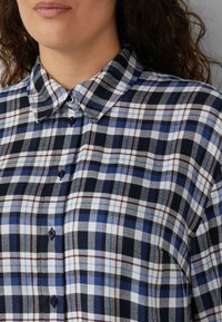 Frau trägt ein blau-weiß-schwarz kariertes Hemd mit Kragen, lockiges dunkles Haar ist auf einer Seite sichtbar, neutraler Hintergrund.