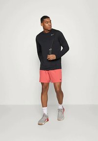 Muškarac u crnoj majici dugih rukava i koraljnim šortsevima drži konopac za skakanje, stojeći nasuprot jednostavne svijetle pozadine.