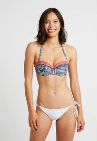 Smilende kvinne i mønstret stroppeløs bikinitopp med halterstropp og hvite sideknute-truser mot en ensfarget bakgrunn.