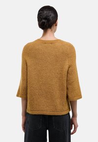 Femme aux cheveux foncés attachés en chignon, portant un pull jaune moutarde à manches courtes et un jean noir, vue de dos sur un fond uni.