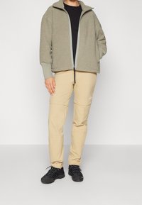 Giacca in pile grigio chiaro con colletto alto e zip, abbinata a pantaloni cargo beige. Scarponi da trekking neri con suole testurizzate.