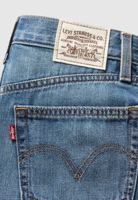 Poche arrière d'un jean Levi's bleu avec coutures orange, étiquette rouge Levi's, et patch beige de la marque représentant deux chevaux et texte.