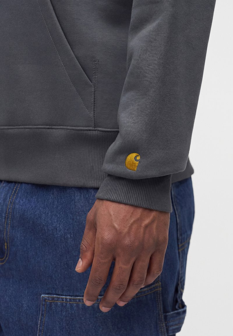 Nahaufnahme einer Person, die ein graues Sweatshirt mit einem gelben Logo am Ärmelbündchen und blaue Jeans mit sichtbaren Nähten trägt.