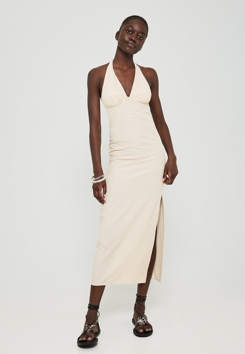 Beiges Halterneck-Kleid mit tiefem V-Ausschnitt, geripptem Textil, figurbetontem Schnitt und seitlichem Schlitz, kombiniert mit schwarzen Riemchensandalen.