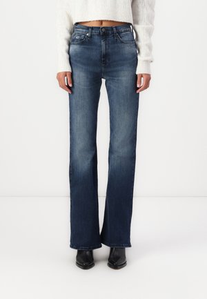 Vrouw draagt een high-waist donkerblauwe flared jeans, een cropped witte trui en zwarte enkellaarsjes, staand tegen een effen witte achtergrond.