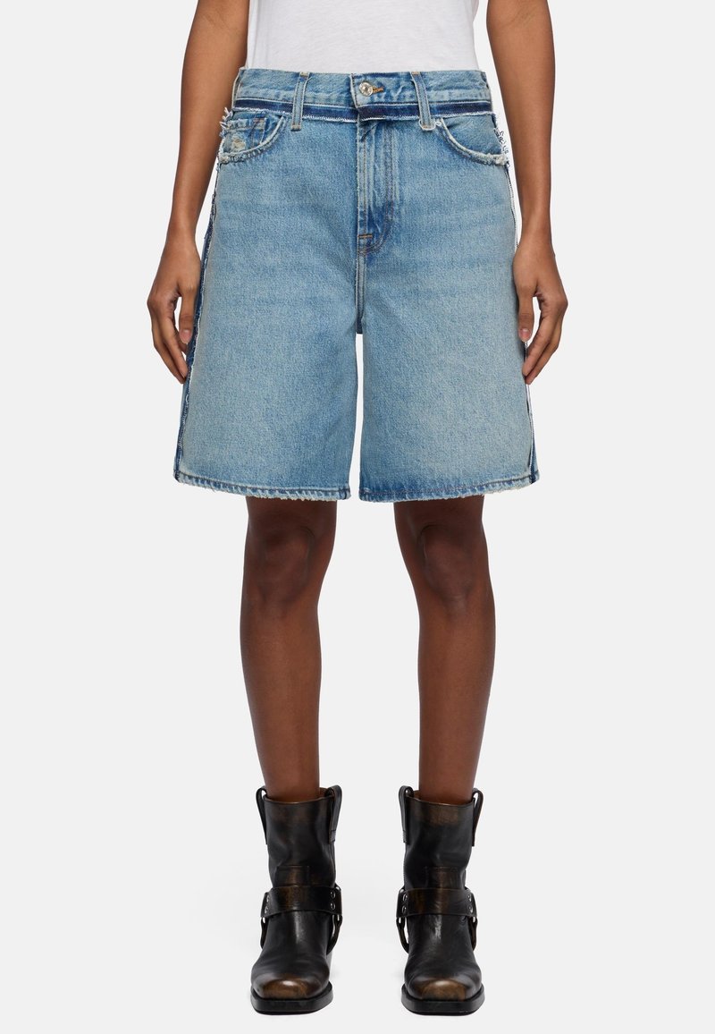 Person iført blå denimshorts til knæene med slidte kanter og sorte læderankelstøvler, stående mod hvid baggrund.