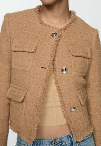 Hnědý tweedový blazer s třepenými okraji, který má dvě přední kapsy a stříbrné knoflíky. Látka má texturovaný povrch.